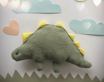 May include: A green and yellow dinosaur plush toy with a textured fabric. The toy is sitting on a green and white background with a yellow sun and blue clouds.