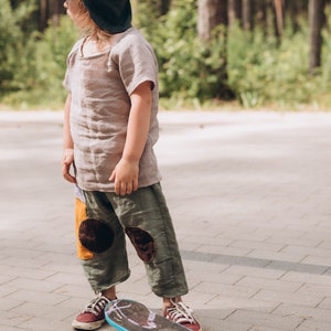 May include: A child in a dinosaur hat and outfit stands on a skateboard. The outfit includes a short-sleeved shirt and olive green trousers with patches. The skateboard has a blue edge and white wheels.