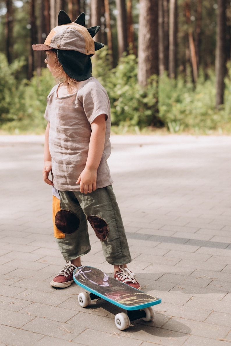 May include: A child in a dinosaur hat and outfit stands on a skateboard. The outfit includes a short-sleeved shirt and olive green trousers with patches. The skateboard has a blue edge and white wheels.