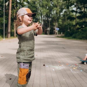 May include: A child in a green linen outfit, including a t-shirt and trousers with colour-block patches, and a dinosaur-themed cap. The child is wearing maroon trainers. The background shows a paved path and trees.
