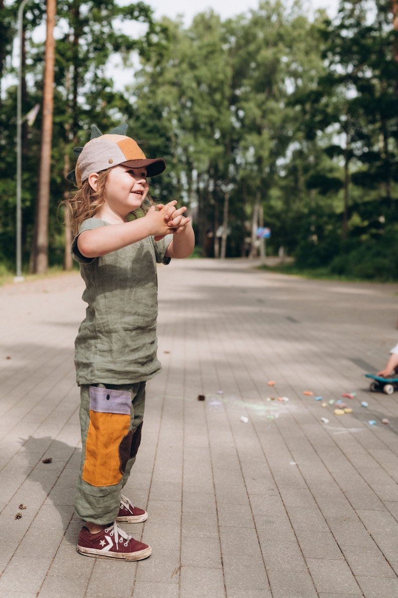 May include: A child in a green linen outfit, including a t-shirt and trousers with colour-block patches, and a dinosaur-themed cap. The child is wearing maroon trainers. The background shows a paved path and trees.