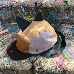 May include: A patchwork cap with a dinosaur-like design. The cap has a brown brim, tan, brown, and grey panels, and dark green spikes. The cap is made of fabric and has a button closure.
