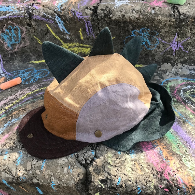 May include: A patchwork cap with a dinosaur-like design. The cap has a brown brim, tan, brown, and grey panels, and dark green spikes. The cap is made of fabric and has a button closure.