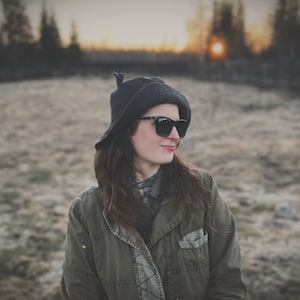May include: A person wearing a dark gray hat, black sunglasses, and an olive green jacket. The person is smiling and looking to the side, with a blurred background of a field and trees.