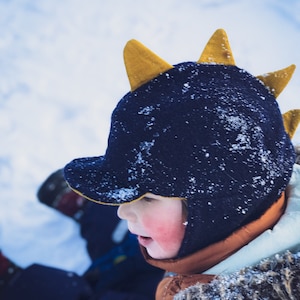Peut inclure: Un enfant portant un bonnet d'hiver bleu et jaune avec un design en pointe. Le bonnet est recouvert de neige et les joues de l'enfant sont roses.