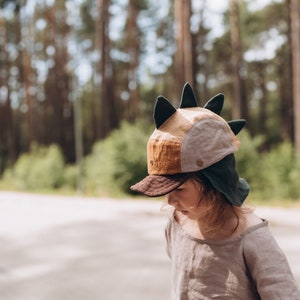 May include: A child's dinosaur-themed hat with a brown peak, tan and brown panels, and dark green details. The hat has black spikes and is worn with a light brown linen top. The background shows a blurred forest.
