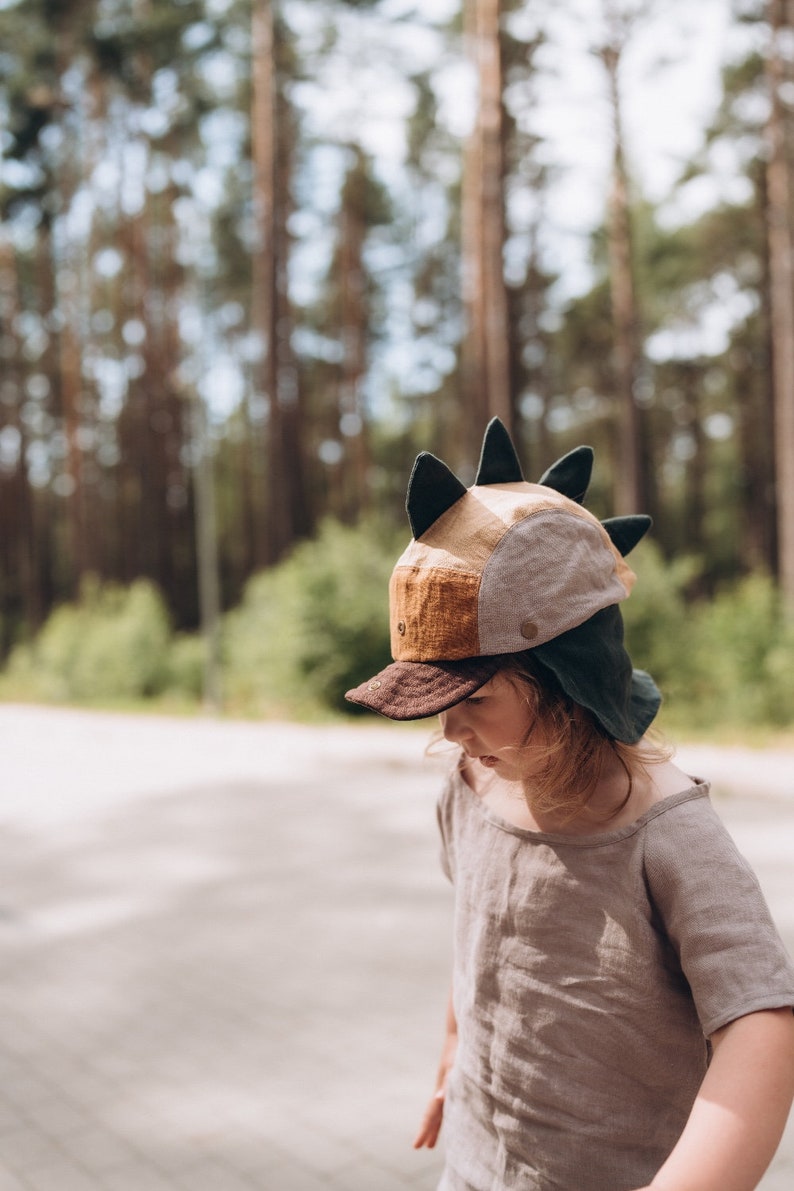 May include: A child's dinosaur-themed hat with a brown peak, tan and brown panels, and dark green details. The hat has black spikes and is worn with a light brown linen top. The background shows a blurred forest.