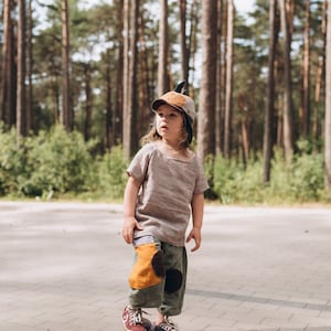 May include: A child wearing a brown cap with a feather, a light brown shirt, and olive green trousers with patches, stands on a skateboard. The child is in a forest setting with tall trees in the background.