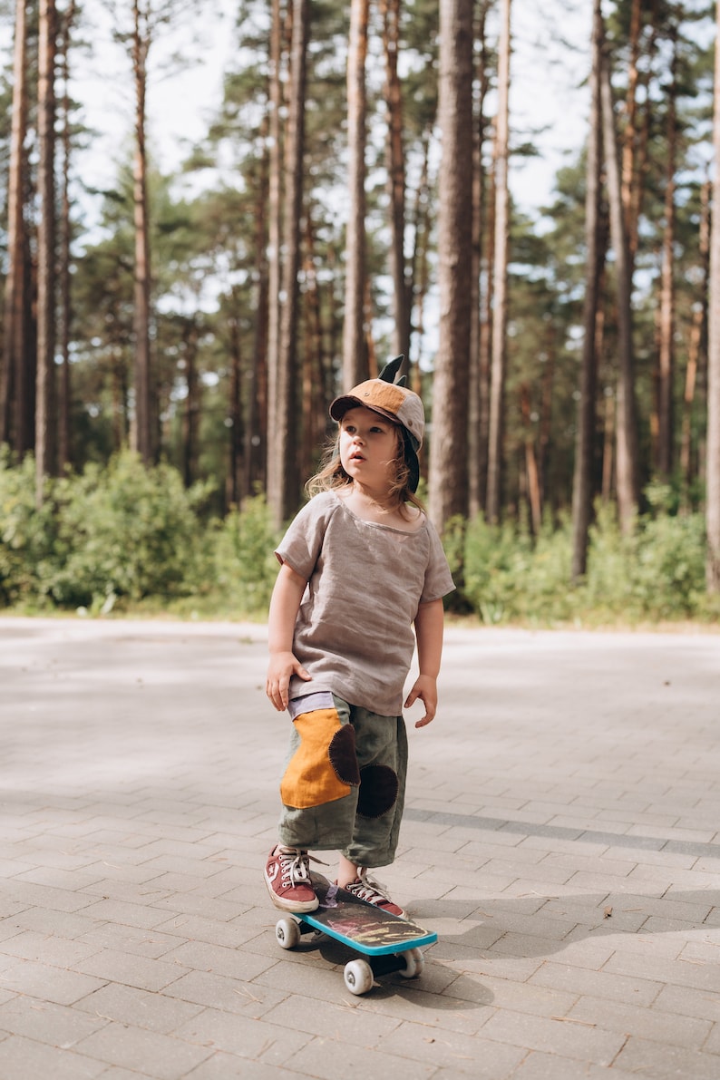 May include: A child wearing a brown cap with a feather, a light brown shirt, and olive green trousers with patches, stands on a skateboard. The child is in a forest setting with tall trees in the background.