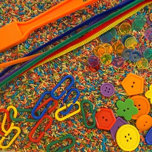 May include: A sensory bin with colourful rice, an orange magnetic wand, pipe cleaners, translucent discs, and various shaped buttons. The buttons are in the shapes of flowers, hexagons, and circles, in shades of orange, yellow, purple, and blue.