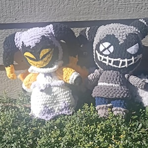 Puede incluir: Dos juguetes de peluche tejidos a crochet. Uno es un bufón morado y blanco con detalles amarillos y cuernos negros. El otro es un monstruo gris y blanco con una amplia sonrisa de dientes y ojos negros.