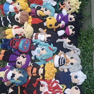May include: A collection of handmade crochet dolls with various hair colors and outfits. The dolls have large, round eyes and are arranged on a bed of green foliage. The dolls are in various colors, including blue, yellow, red, and black.