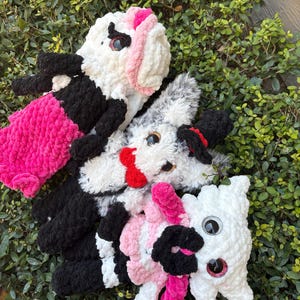 May include: A collection of handmade crochet stuffed animals in various colors. The toys include a white and black cow, a white and pink bunny, and a gray and black dog. The toys have large, googly eyes and are set against a backdrop of green foliage.