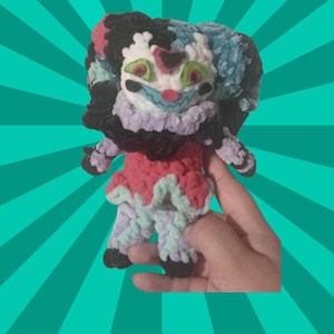 May include: A handmade plush doll with a clown-like design. The doll has a white face with green eyes, a blue mouth, and a heart-shaped detail. It has black, blue, and white hair, a ruffled collar, and a red and pink outfit. The doll is held against a teal background.