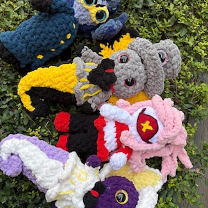 May include: A collection of colorful, handmade crochet creatures in various shapes and sizes. The plush toys feature vibrant colors like blue, yellow, pink, and purple, with detailed eyes and whimsical designs. These soft sculptures are arranged on a bed of green foliage.