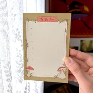 以下が含まれることがあります： ベージュの背景と薄茶色の縁取りのある、長方形の To Do List ノートパッド。 ノートパッドの上部には、「To Do List」という言葉が入ったピンク色のバナーがあります。 ノートパッドには、キノコとブドウのイラストが描かれています。