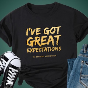 May include: Black t-shirt with gold lettering that reads "I've Got Great Expectations" and "The Outsiders A New Musical".
