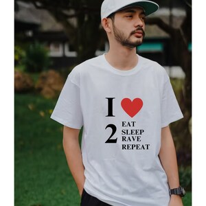 Puede incluir: Camiseta blanca con un gráfico negro que dice "I 2 EAT SLEEP RAVE REPEAT" con un corazón rojo reemplazando el "love" de la frase.