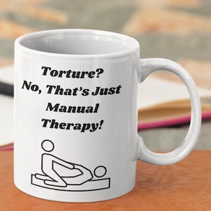 Puede incluir: Taza de cerámica blanca con el texto "Torture? No, That's Just Manual Therapy!" en negro. Debajo del texto hay una ilustración en blanco y negro de una persona recibiendo un masaje. La taza tiene un asa curvada.