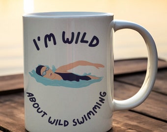 May include: White ceramic mug with the text "I'M WILD ABOUT WILD SWIMMING" in navy blue. The mug features an illustration of a swimmer in a black swimsuit and goggles, swimming in blue water. The mug has a curved handle.