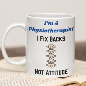 Puede incluir: Taza de cerámica blanca con el texto "I'm A Physiotherapist I Fix Backs NOT ATTITUDE" en azul y negro. Un gráfico de columna vertebral aparece debajo de "I Fix Backs". La taza tiene un asa curva y está sobre un libro.