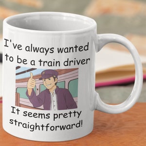 Pode incluir: Caneca de cerâmica branca com uma ilustração em preto e branco de um maquinista de trem dando um polegar para cima. O texto na caneca diz "Eu sempre quis ser maquinista. Parece bem simples!"