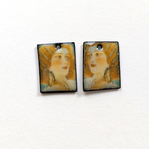 May include: Two rectangular pendants with a vintage portrait of a woman with long, flowing, reddish-blonde hair. The portraits are framed with a black border and have a small hole at the top for hanging. The background is a light cream color.