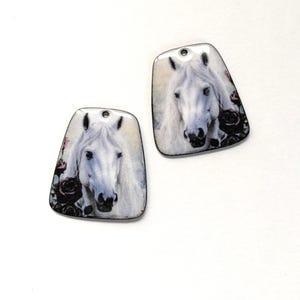 May include: Two trapezoidal pendants featuring a detailed illustration of a white horse with a flowing mane, accented by dark roses. Each pendant has a smooth, glossy finish and a small hole at the top for attachment to a necklace or cord.