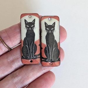 Puede incluir: Dos colgantes rectangulares con una ilustración de un gato negro centrada sobre un fondo circular de color crema y naranja. Cada colgante tiene un acabado liso y dos pequeños orificios circulares, uno arriba y otro abajo, para sujeción.