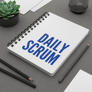 May include: A white spiral bound notebook with the words "DAILY SCRUM" written in blue letters. The notebook is on a gray surface with a pencil and a plant in a pot.