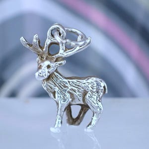 May include: A silver charm depicting a deer with detailed antlers and textured body. The charm has a loop for attaching to jewellery. The deer is standing on a white surface. The background is out of focus.