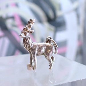 May include: A silver deer charm with detailed antlers and body. The charm is standing on a white surface, with a blurred background of pink, grey, and white. The charm has a small loop at the top for attaching to a necklace or bracelet.