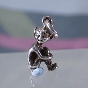 Op de afbeelding: Een zilverkleurige bedel die een zittend, grillig figuur met puntoren en een kleine hoed afbeeldt. Het figuur omarmt zijn knieën. De bedel heeft een lus om aan een ketting of armband te bevestigen. De basis van de bedel heeft een lichtblauwe kleur.