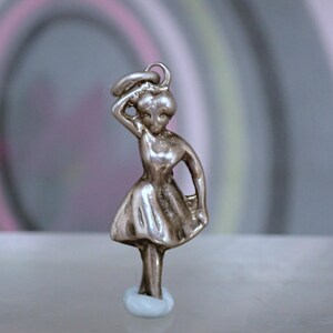 May include: A metallic silver-toned charm shaped like a figure wearing a dress with a flared skirt. The figure has one arm raised to the head and the other arm positioned at the side. A small circular loop is attached to the top of the head.