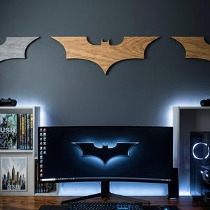 May include: A computer setup featuring a large curved monitor displaying the Batman logo. Wooden Batman symbols are mounted on the dark gray wall above. The desk includes a keyboard, mouse, and a black mousepad. Shelves with books and a Gotham poster are on the left.