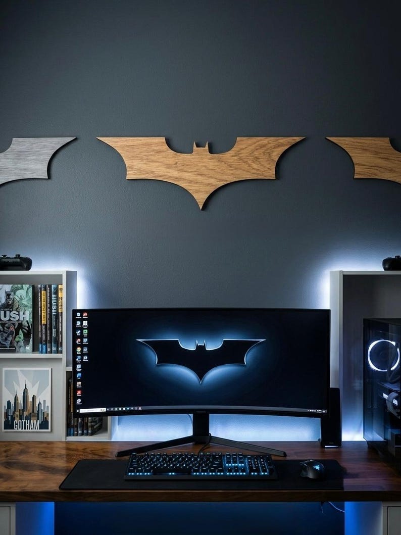 May include: A computer setup featuring a large curved monitor displaying the Batman logo. Wooden Batman symbols are mounted on the dark gray wall above. The desk includes a keyboard, mouse, and a black mousepad. Shelves with books and a Gotham poster are on the left.