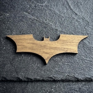 May include: Wooden Batman logo in a brown finish. The iconic bat-shaped emblem is displayed against a dark, textured background. This decorative item is suitable for fans of comic books and superhero themes.