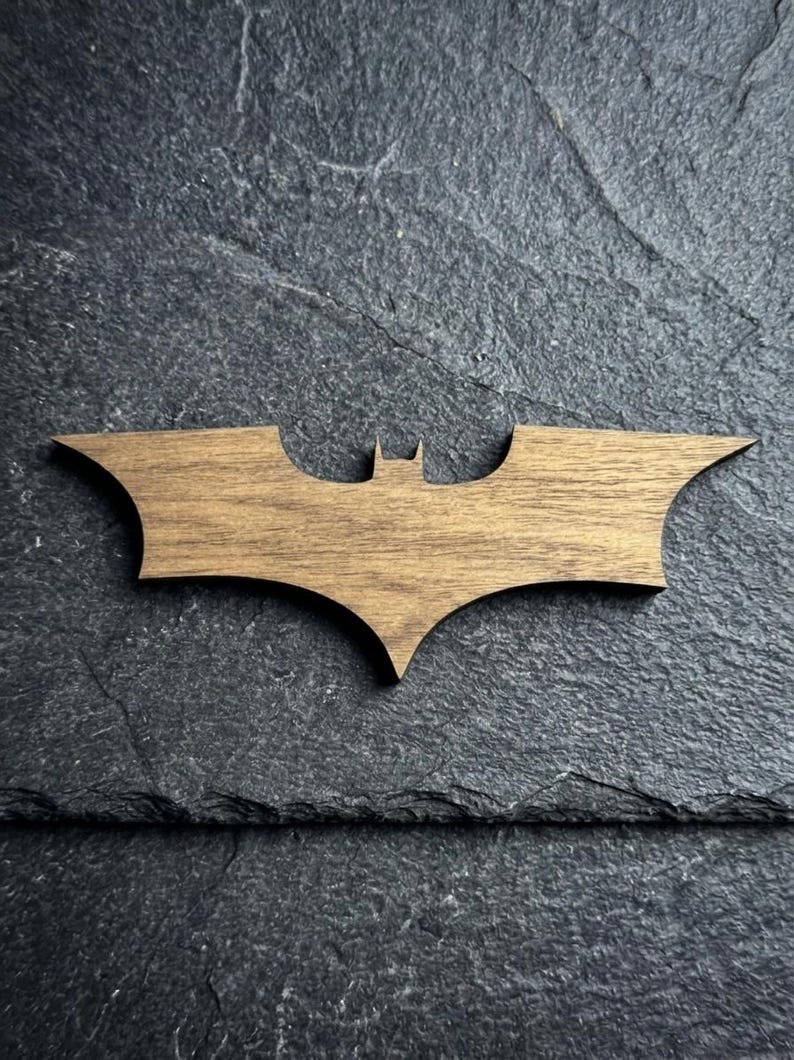 May include: Wooden Batman logo in a brown finish. The iconic bat-shaped emblem is displayed against a dark, textured background. This decorative item is suitable for fans of comic books and superhero themes.