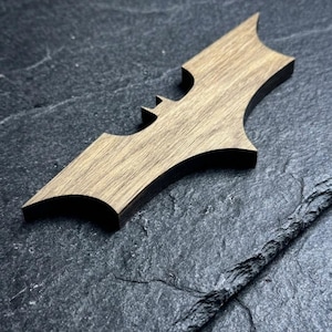 May include: A wooden Batman logo in a batarang shape, with a light brown wood grain. The bat-shaped emblem is set against a dark, textured background. The design is a stylized representation of the iconic superhero symbol.