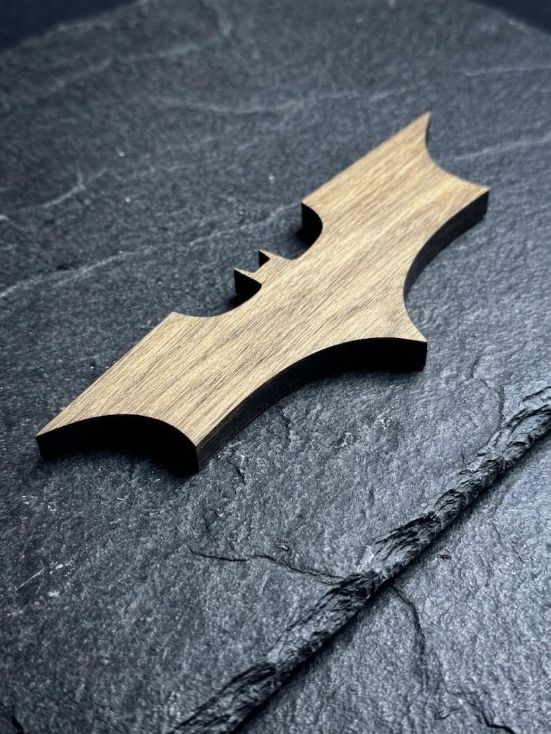 May include: A wooden Batman logo in a batarang shape, with a light brown wood grain. The bat-shaped emblem is set against a dark, textured background. The design is a stylized representation of the iconic superhero symbol.