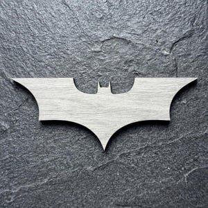 May include: A light gray wooden Batman logo, featuring the iconic bat silhouette. The emblem is set against a dark gray, textured background. The design is a stylized representation of the superhero's emblem.