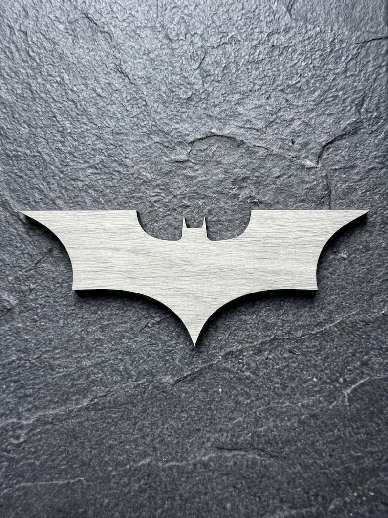 May include: A light gray wooden Batman logo, featuring the iconic bat silhouette. The emblem is set against a dark gray, textured background. The design is a stylized representation of the superhero's emblem.