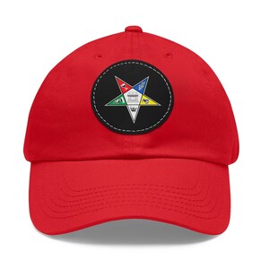 Puede incluir: Gorra de béisbol roja con un parche negro y blanco que presenta una estrella de cinco puntas con un símbolo masónico en el centro.