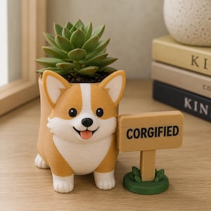 Puede incluir: Una maceta con forma de corgi en tonos beige y blanco que contiene una suculenta verde. La maceta muestra una cara de corgi sonriente con una lengua rosa. Junto a la maceta hay un pequeño letrero marrón claro sobre una base verde con el texto CORGIFIED.