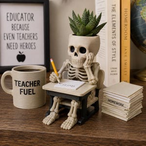 May include: A beige skeleton figurine sitting at a small desk, holding a yellow pencil over a white paper labeled Quiz. The skeleton's head serves as a planter for a green succulent. Beside it sits a beige mug with Teacher Fuel printed in black.