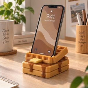 May include: A smartphone resting on a waffle-shaped stand. The stand features two stacked golden-brown waffles with a square pat of yellow butter on top and brown syrup dripping down the sides. The phone screen displays the time 9:41.