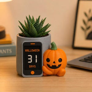 May include: A gray cylindrical planter containing a green succulent, featuring a digital display shaped like a smartphone that reads HALLOWEEN 31 DAYS in orange and white text. Beside it sits a small, orange, smiling jack-o'-lantern figurine.