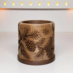 May include: A cylindrical brown candle holder with a textured wood-like finish. The exterior features an embossed design of pine branches, pinecones, and small snowflakes. The item sits on a flat, circular base of a slightly darker brown shade.