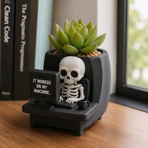 May include: A dark grey planter featuring a white skeleton figurine sitting at a computer desk. The computer screen displays the text IT WORKED ON MY MACHINE. A green succulent plant is potted in the top section of the planter, which also includes a small black mug on the desk surface.