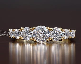 May include: A gold band ring featuring seven round-cut diamonds. The diamonds are set closely together, creating a continuous sparkle. The ring is displayed on a dark, reflective surface, enhancing its brilliance. The text "Brilliant Karat" is visible.